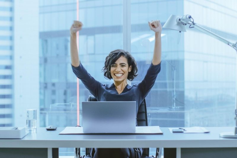 woman sitting at a desk with a laptop in front of her celebrating with her arms in the air